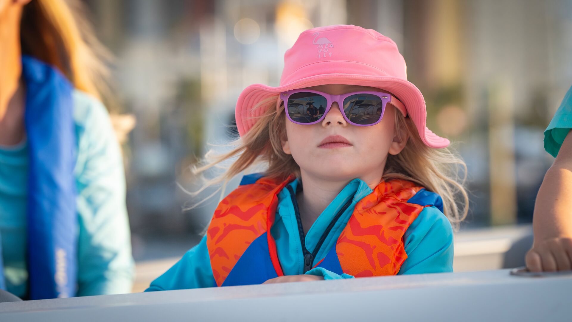 A young girl in a life jacket and a hat with sunglasses. 