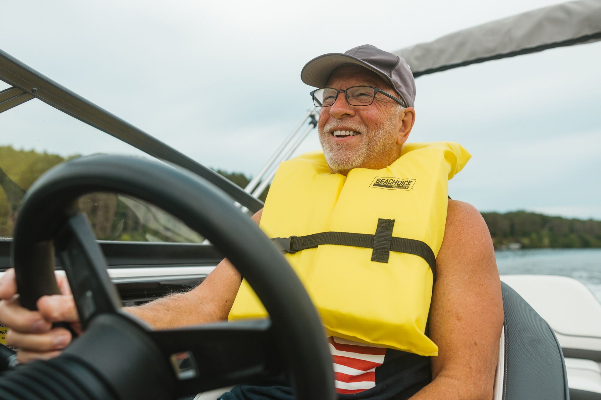 A man smiles while driving a boat and wearing a yellow life vest. 